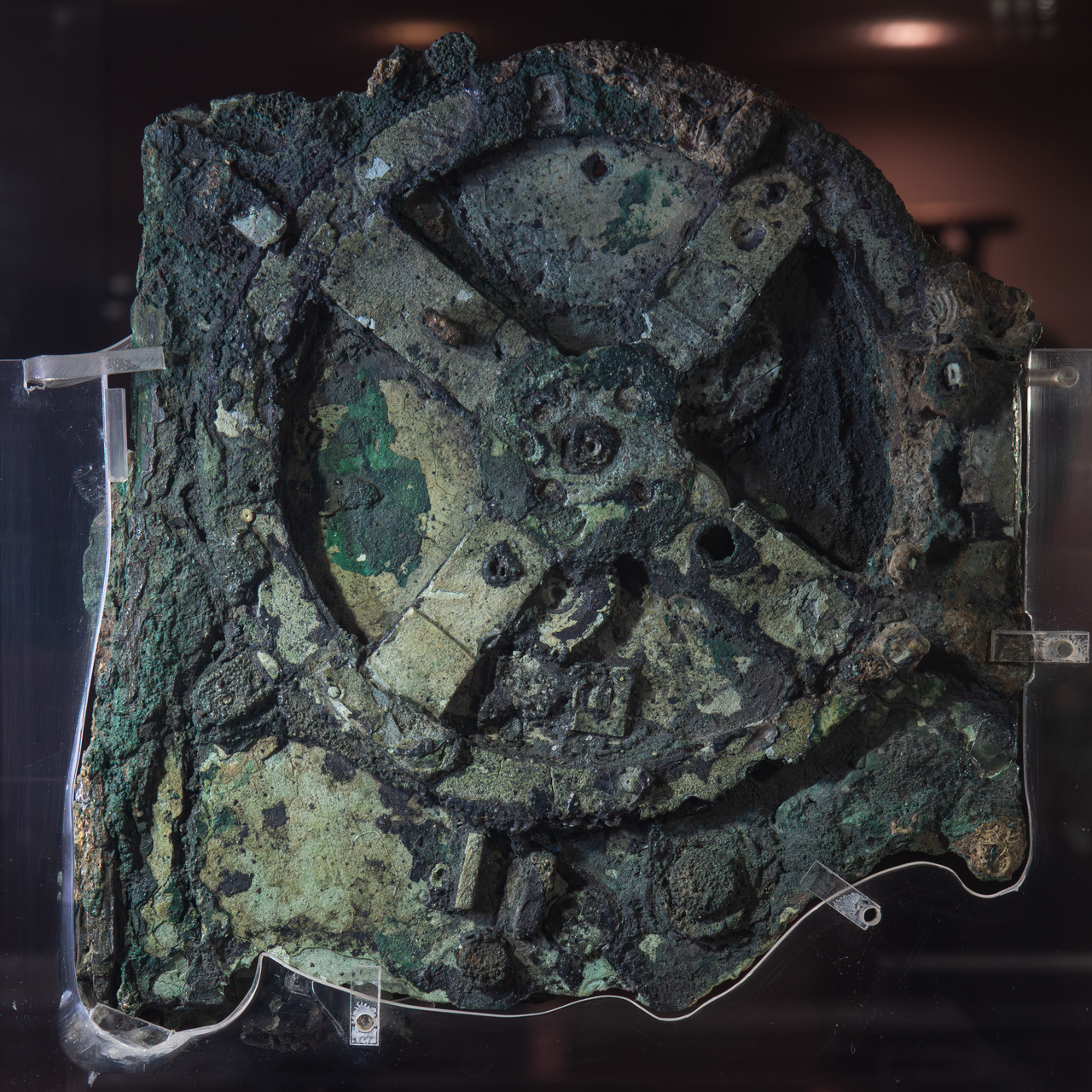 A heavily oxidized, ancient bronze gear mechanism set within weathered rock or calcified material. The main fragment shows a large, complex geared wheel with smaller gears visible, indicating intricate, ancient mechanical engineering.